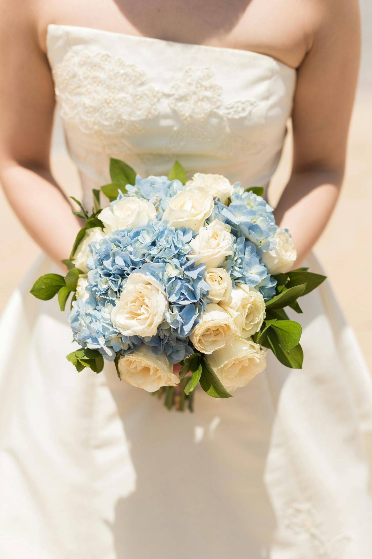 Bride holding a bouquet of blue hydrangeas and white roses against a white wedding dress.