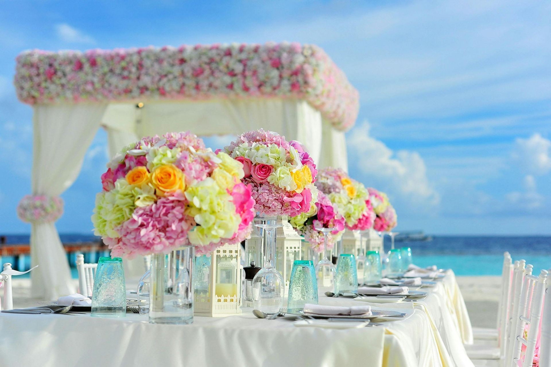 Elegant beach wedding table setup with colorful floral centerpieces under a decorated canopy.